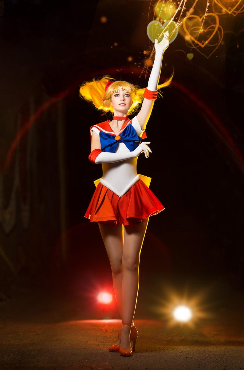May include: A woman dressed as a Sailor Moon character with a red and orange skirt, white top, and blue bow. She is holding her hands up in the air, and there are glowing hearts around her.