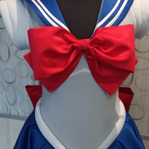 May include: A blue and white sailor-style dress with a large red bow. The dress has a white collar and a blue skirt with a white trim.