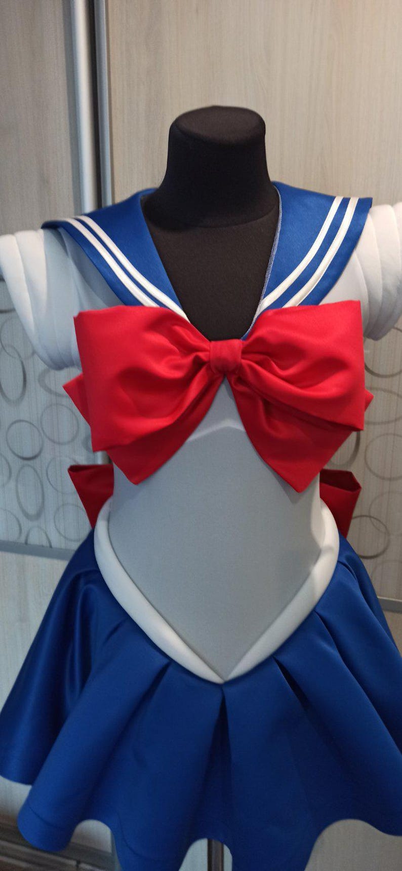 May include: A blue and white sailor-style dress with a large red bow. The dress has a white collar and a blue skirt with a white trim.
