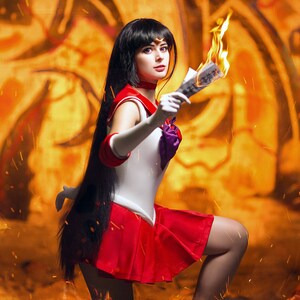 May include: A woman dressed as a Sailor Moon character with long black hair and a red and white costume. She is holding a burning piece of paper in her right hand and is looking directly at the camera.