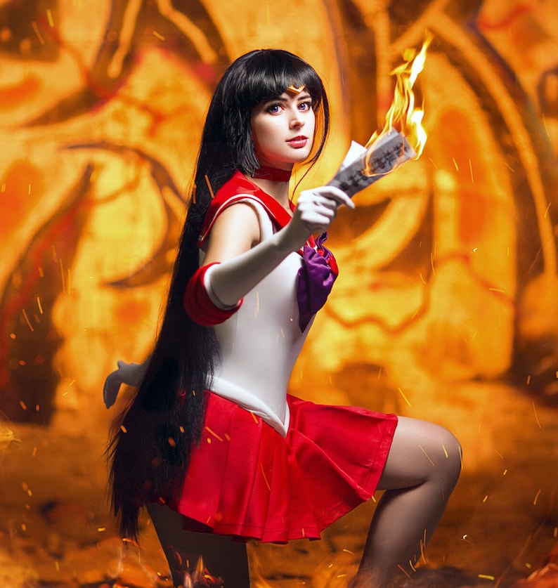 May include: A woman dressed as a Sailor Moon character with long black hair and a red and white costume. She is holding a burning piece of paper in her right hand and is looking directly at the camera.