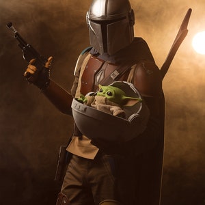 May include: A Mandalorian in a silver helmet holds a small green creature in a round, gray pod. The Mandalorian is wearing brown and tan armor and a dark cape. The background is a hazy brown.