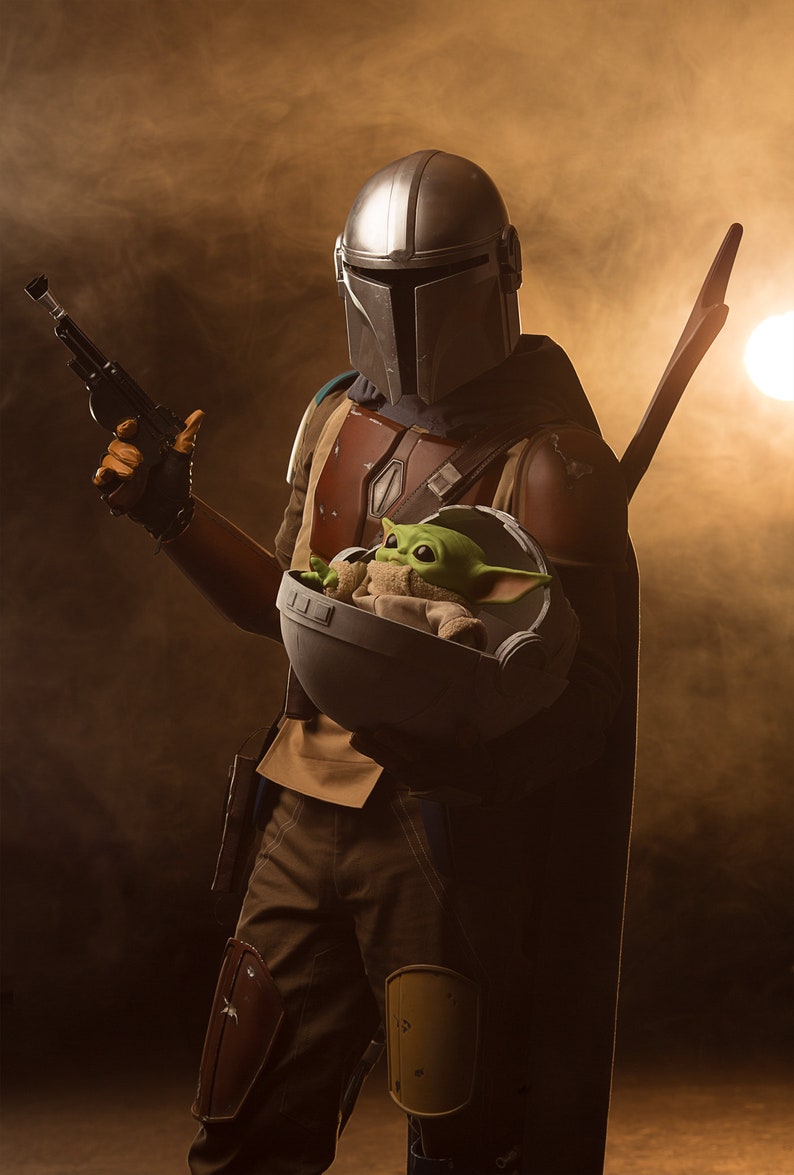 May include: A Mandalorian in a silver helmet holds a small green creature in a round, gray pod. The Mandalorian is wearing brown and tan armor and a dark cape. The background is a hazy brown.