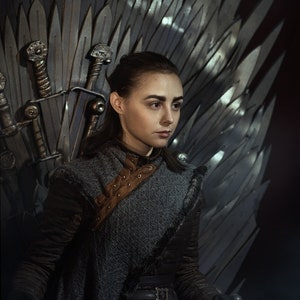 Puede incluir: Una mujer con una túnica gris oscura y una correa de cuero marrón se sienta en un trono hecho de espadas. Lleva un cinturón y guantes de cuero negro.