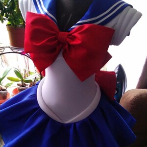 May include: A white sailor-style dress with a blue collar and red bow. The dress has a blue skirt with a ruffled hem.