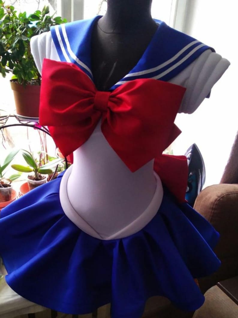 May include: A white sailor-style dress with a blue collar and red bow. The dress has a blue skirt with a ruffled hem.