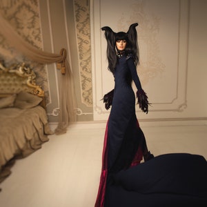 May include: A woman in a long black dress with a red train and black feathered accents stands in a room with a large bed and a side table. She has long black hair with two black feathers on top of her head.