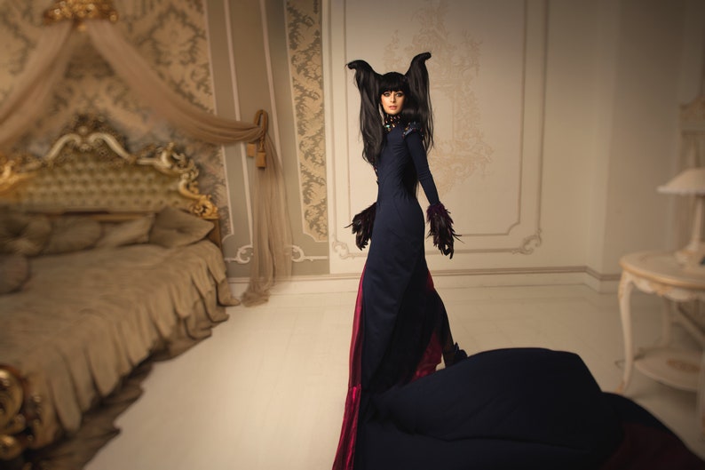 May include: A woman in a long black dress with a red train and black feathered accents stands in a room with a large bed and a side table. She has long black hair with two black feathers on top of her head.