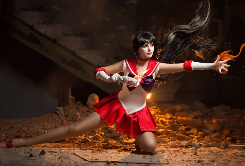 May include: A woman dressed as a Sailor Moon character with long black hair and a red and white costume. She is holding a glowing object in her right hand and her left hand is outstretched. She is standing in a dark, industrial setting with rubble and debris in the background.