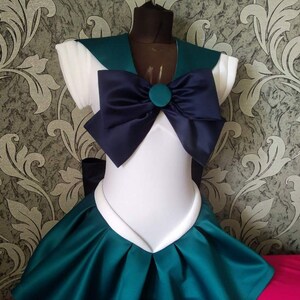 May include: A white and teal sailor-style dress with a large navy blue bow and a teal button. The dress has a white collar and short sleeves.