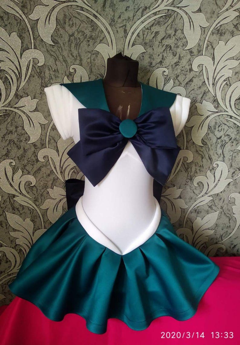 May include: A white and teal sailor-style dress with a large navy blue bow and a teal button. The dress has a white collar and short sleeves.
