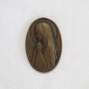 May include: Oval-shaped bronze religious pendant featuring a woman in profile with hands clasped in prayer. The woman is wearing a draped head covering and is set against a halo. The pendant has a detailed, embossed design.