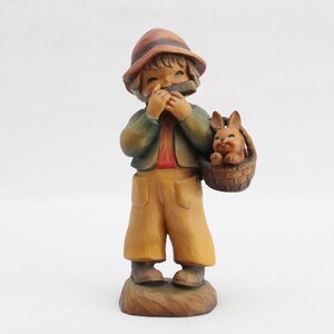 May include: A wooden figurine of a boy wearing a brown hat and green jacket, playing a harmonica. He is holding a basket with a small rabbit inside.