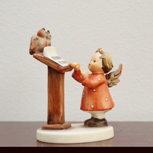 May include: A ceramic figurine of a young angel in a red dress with white dots, wings, and brown shoes. The angel is conducting two birds perched on a wooden music stand with sheet music. The figurine is on a white oval base.