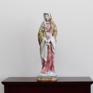 May include: A ceramic statue of a woman in a pink and yellow robe, holding a green branch. The statue is standing on a pedestal with a floral design.