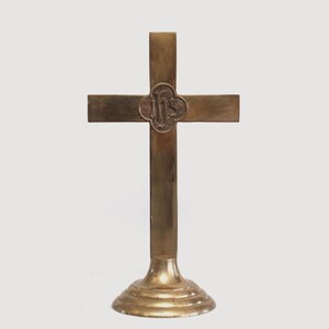 May include: A gold-colored metal cross with a decorative design on the front. The cross is mounted on a round base.