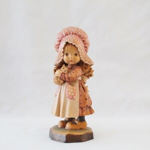 May include: A wooden figurine of a girl wearing a pink and white polka dot dress and a pink and white polka dot bonnet. She is holding a small brown and white kitten in her arms. The figurine is standing on a wooden base.