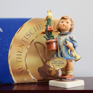 May include: A Hummel figurine of a young girl in a blue dress with red polka dots, holding a trumpet and a potted plant with a yellow bird. The figurine has a gold tag that reads "FINAL ISSUE LETZTE AUSGABE 1999".