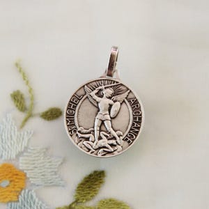 May include: A silver pendant featuring St. Michael the Archangel. The round pendant depicts St. Michael with a sword and shield, standing over figures. The words "ST. MICHEL ARCHANGE" are inscribed around the edge. The pendant has a loop for a chain.
