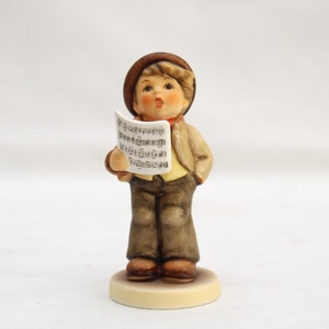 May include: A porcelain figurine of a boy wearing a brown hat and brown pants, holding a sheet of music and singing.
