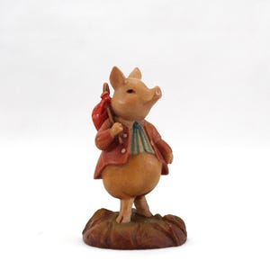 May include: A whimsical figurine of a pig wearing a red jacket and carrying a red sack over its shoulder. The pig is standing on a brown base, with a green and white striped shirt. The figurine is made of a light brown material.
