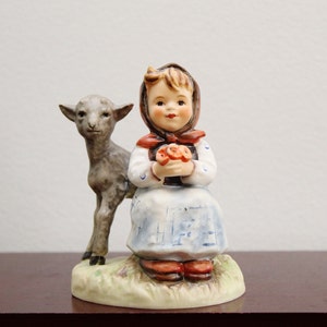 May include: A porcelain figurine of a young girl in a blue and white dress holding a bouquet of flowers. She is standing next to a small gray lamb.