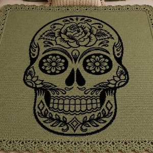 May include: A square crochet blanket in sage green featuring a large, centered black skull design with intricate floral and leaf patterns. The blanket has a decorative scalloped border and a flat, textured stitch throughout the main body.