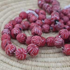 May include: Strands of red and white patterned beads. The beads are round and have a swirl design. The beads are strung together on a white string. The beads are resting on a woven, circular surface.