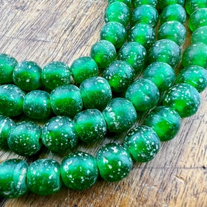 May include: Close-up of strands of green glass beads. The beads are round, translucent, and have a frosted finish with white speckles. They are displayed on a wooden surface, suitable for jewellery making.