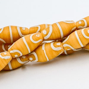 May include: Close-up of a strand of orange-yellow beads with white, curved stripe designs. The beads are oval-shaped and appear to be made of a solid material. The beads are strung together, creating a necklace or bracelet.