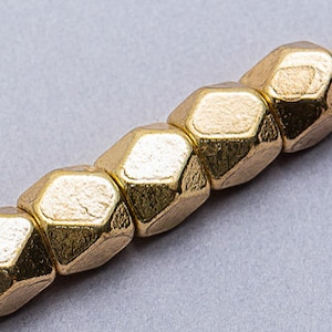 May include: A strand of gold-colored metal beads with a faceted, hexagonal shape. The beads are strung on a thin, beige cord.