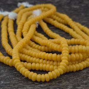May include: Close-up of several strands of yellow beads. The beads are small, round, and have a slightly ribbed texture. The strands are intertwined, with white string visible at the ends. The beads have a warm, sunny color.