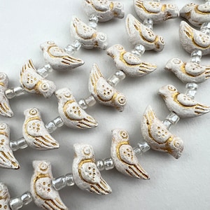 May include: A strand of white ceramic bird beads with gold accents. The beads are strung on a white thread with clear beads in between.