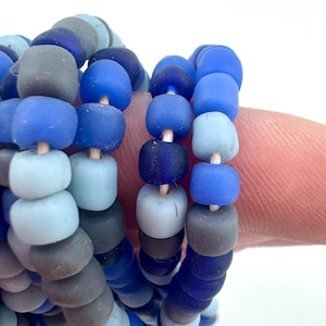 May include: Close-up of several strands of blue glass beads in varying shades, including light blue, medium blue, and dark blue. The beads are cylindrical and strung together, creating a layered effect. The beads are on a white background.