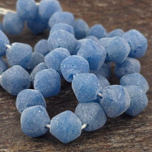 May include: A close-up of a strand of light blue, faceted beads. The beads have a textured, matte finish and are strung on a white cord. The beads are irregularly shaped and appear to be made of glass or a similar material.