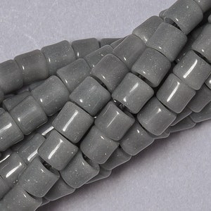 May include: A strand of gray glass beads with a slightly speckled finish. The beads are cylindrical and have a slightly rounded shape.