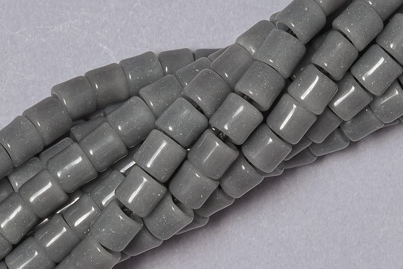May include: A strand of gray glass beads with a slightly speckled finish. The beads are cylindrical and have a slightly rounded shape.