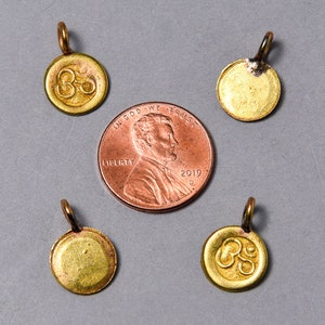 May include: Four gold-colored metal charms with a circular shape. Three of the charms have the Om symbol engraved on them. The fourth charm is plain. The charms are arranged around a US penny.