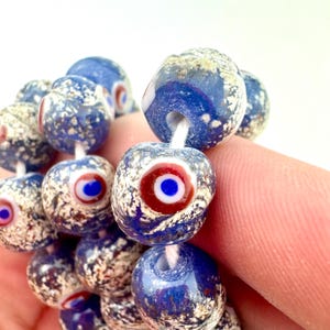 May include: Close-up of a strand of blue and white glass beads. Each bead has a speckled pattern and a central eye design with red and blue accents. The beads are strung on a white cord.