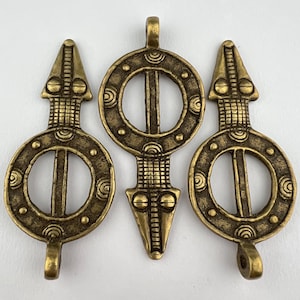 May include: Three gold-toned metal pendants with a circular design and a pointed top. Each pendant features a circle with a smaller circle inside, and a line running through the center. The pointed top of each pendant has a textured design.