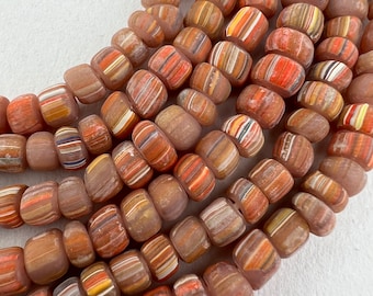 May include: A strand of colourful glass beads with a layered, striped pattern in shades of brown, orange, and white. The beads are opaque and have a slightly frosted finish.