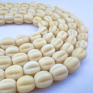May include: Cream-colored, oval-shaped beads with three horizontal grooves. The beads are arranged in a curved pattern, suggesting a necklace or bracelet design. The beads have a smooth, matte finish.