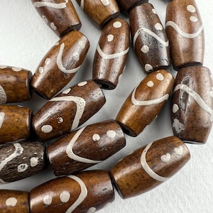 May include: A collection of brown wooden beads with white painted designs. The beads are oval shaped and have a variety of patterns, including dots, lines, and swirls.