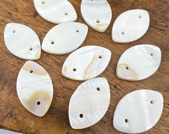 May include: Ten white oval-shaped shell buttons with two holes each. The buttons have a pearlescent sheen and are arranged in a random pattern on a wooden surface.