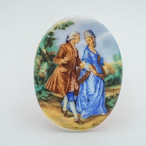 May include: An oval ceramic tile with a painted scene of a couple in 18th-century clothing walking in a garden. The man is wearing a brown coat and the woman is wearing a blue dress.