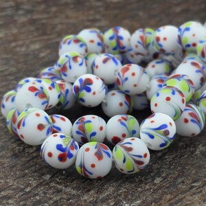 May include: A strand of white glass beads with colorful designs. Each bead features a pattern of red dots, blue swirls, and green and yellow accents. The beads are round and have a matte finish, perfect for jewelry making.