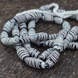 May include: A strand of oval-shaped beads with a black, white, and gray striped pattern. The beads are strung together with a white cord. The beads have a unique design, with curved stripes that create a feather-like effect. The beads are on a dark wooden surface.