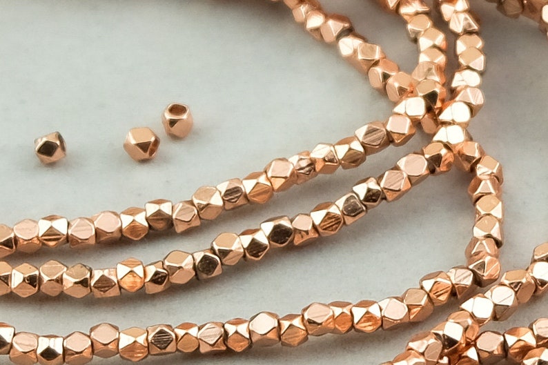May include: A strand of rose gold faceted beads. The beads are small and have a geometric shape. There are also three loose beads in the image.