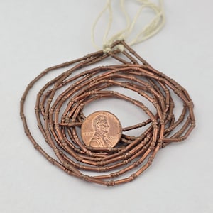 May include: A strand of metallic copper-colored tube beads with small raised rings, arranged in a circular coil. A copper-colored United States one-cent coin is placed in the center for scale, showing the profile of Abraham Lincoln and the year 2006.