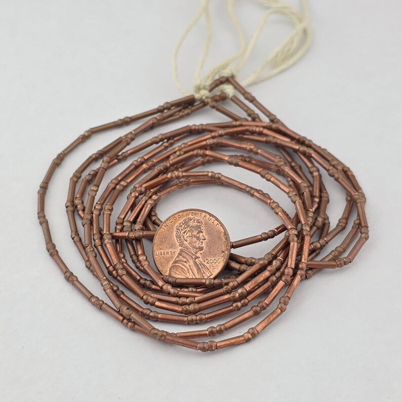 May include: A strand of metallic copper-colored tube beads with small raised rings, arranged in a circular coil. A copper-colored United States one-cent coin is placed in the center for scale, showing the profile of Abraham Lincoln and the year 2006.